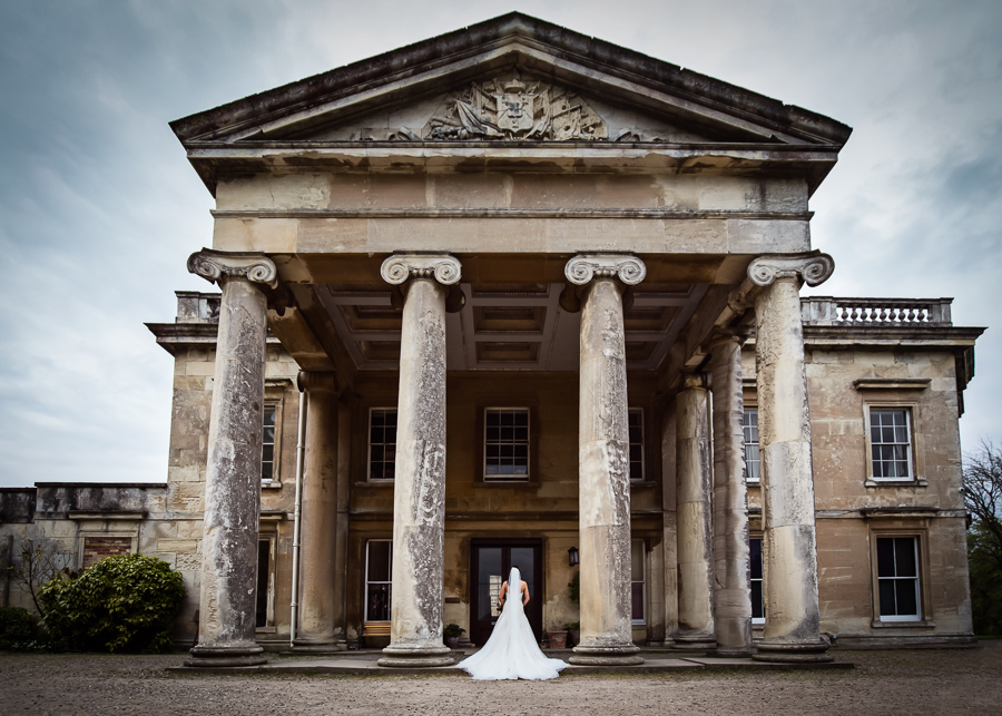 Bride standing alone in front of a grand neoclassical mansion with imposing columns.