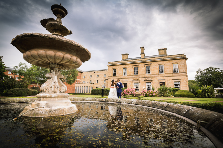 A couple holding hands near a fountain in the foreground, with an elegant mansion in the background.