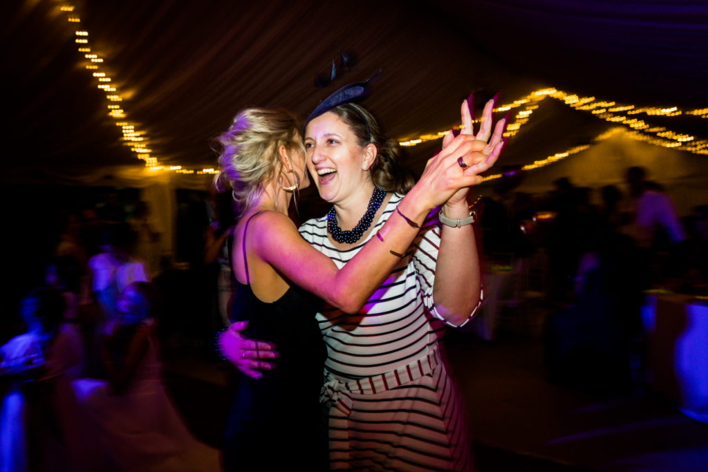 Two women joyfully dancing together at an event with a tent and string lights in the background.