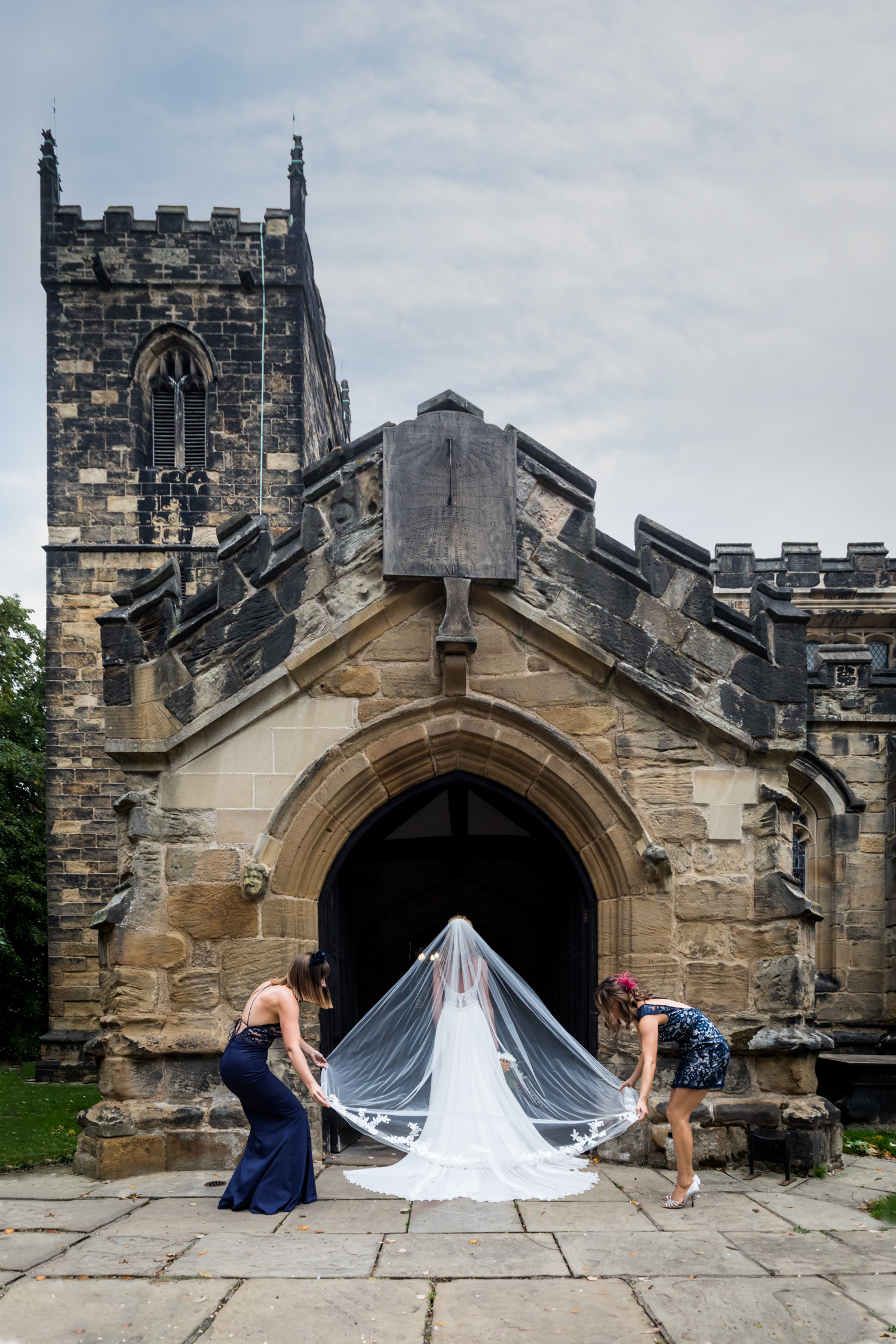 Two people adjusting a bride's long veil in front of a church entrance.
