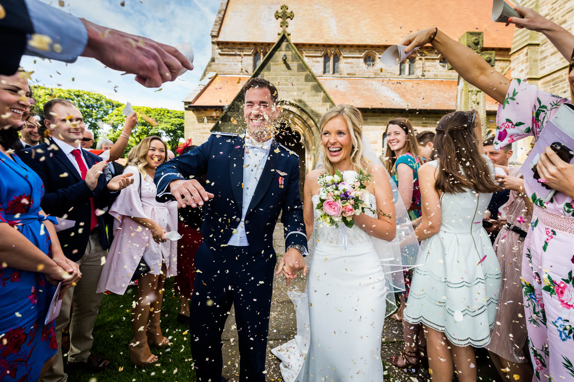 Bride and groom smiling while walking through a shower of confetti with guests celebrating around them.