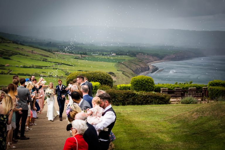Couple walking through confetti tunnel in front of coastal view at Raven Hall