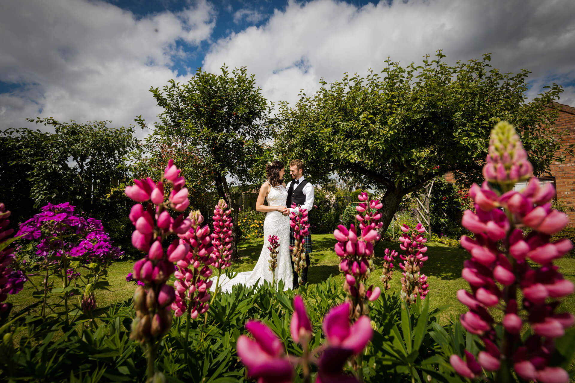 A couple stands embracing in a garden, surrounded by vibrant flowers and greenery under a cloudy sky.