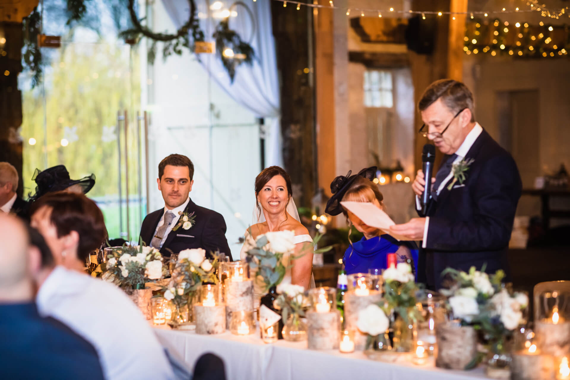 A wedding reception scene with a couple seated at a table adorned with floral arrangements and candles, attentively listening to a man giving a speech.