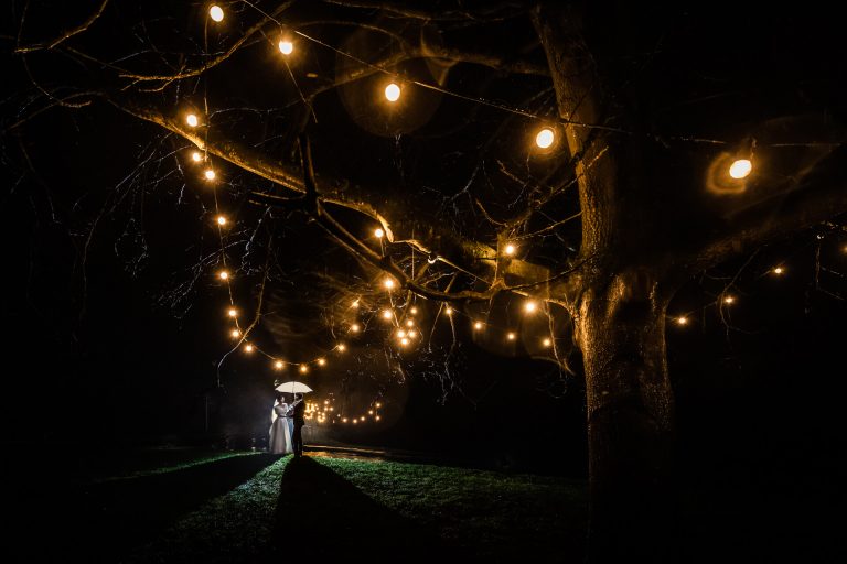 A couple stands beneath a tree adorned with glowing lanterns at night, casting a warm light on the surroundings.