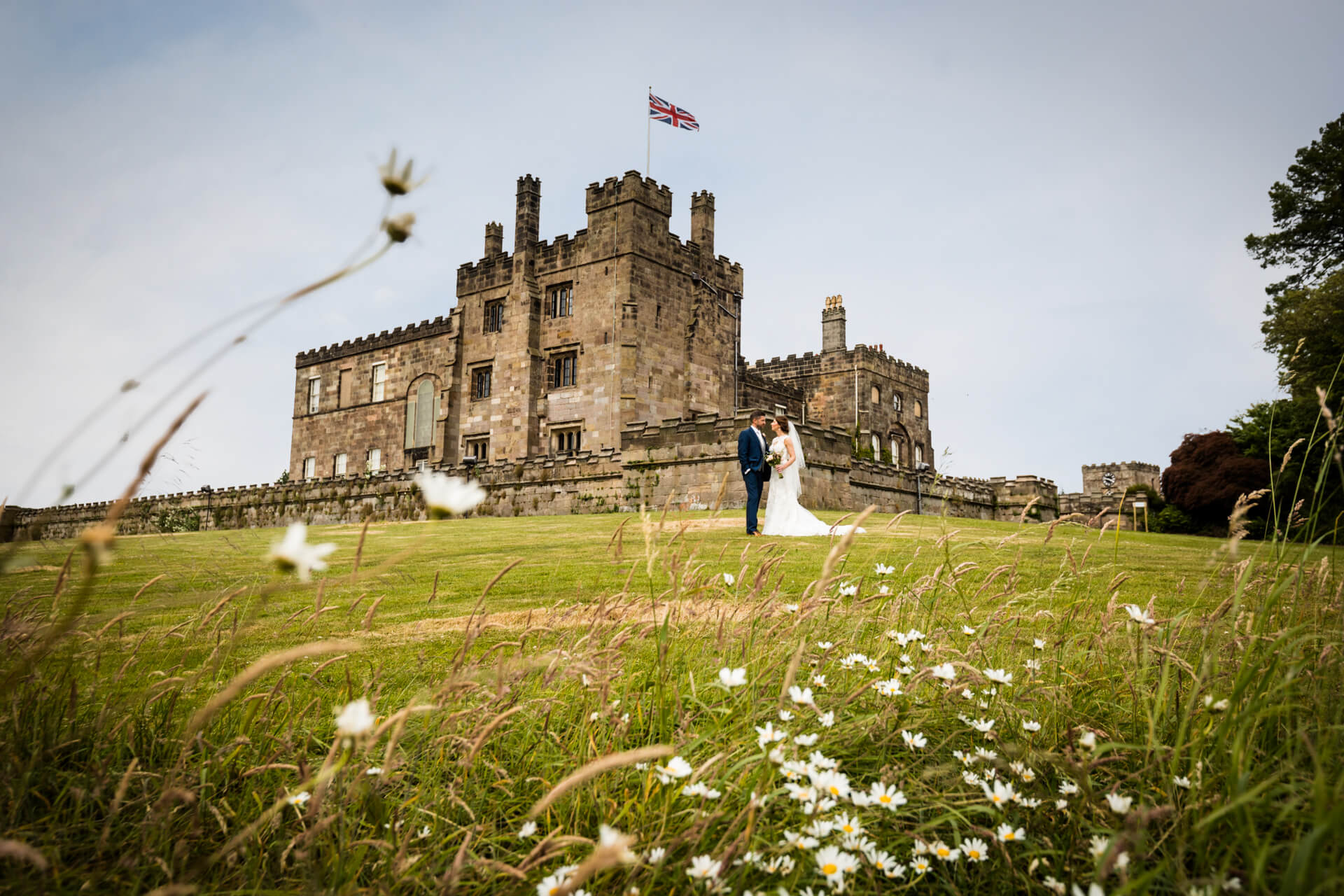 A bride and groom holding hands in front of a grand castle with a flag flying atop under a cloudy sky, with wildflowers in the foreground.