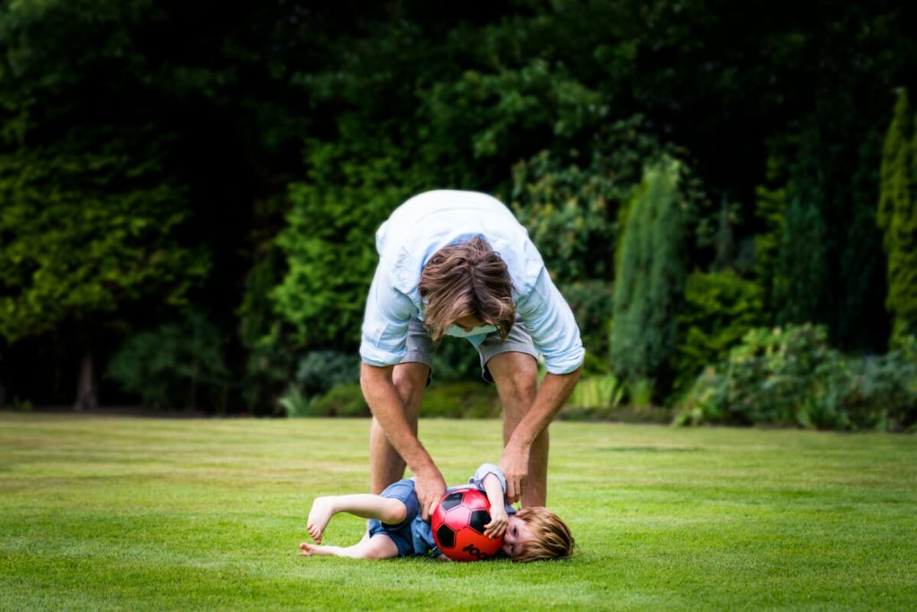 dad tickling his son as he lies on the grass clothing his football