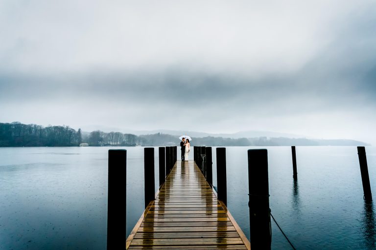 A person standing at the end of a wet wooden pier extending into a tranquil lake under an overcast sky.