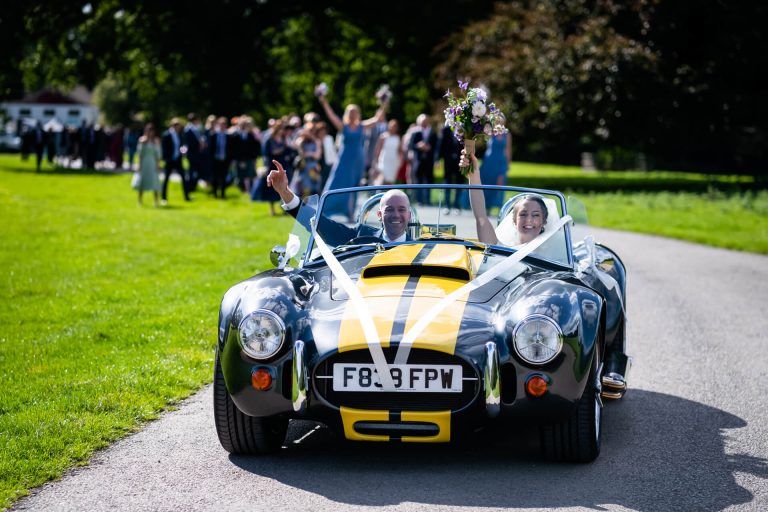 bride waves her bouquet from the wedding car and guests walk behind