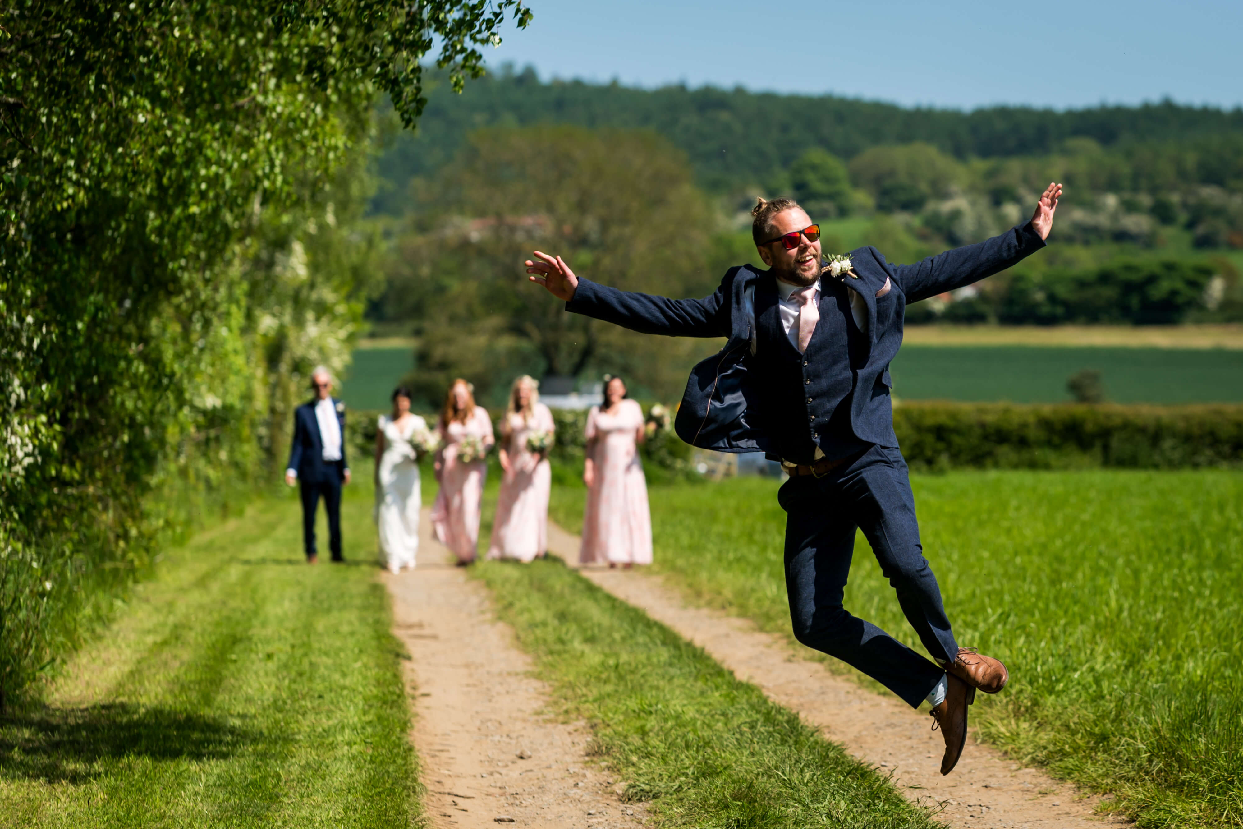 Groomsman joyfully jumping, bridal party walking in countryside.