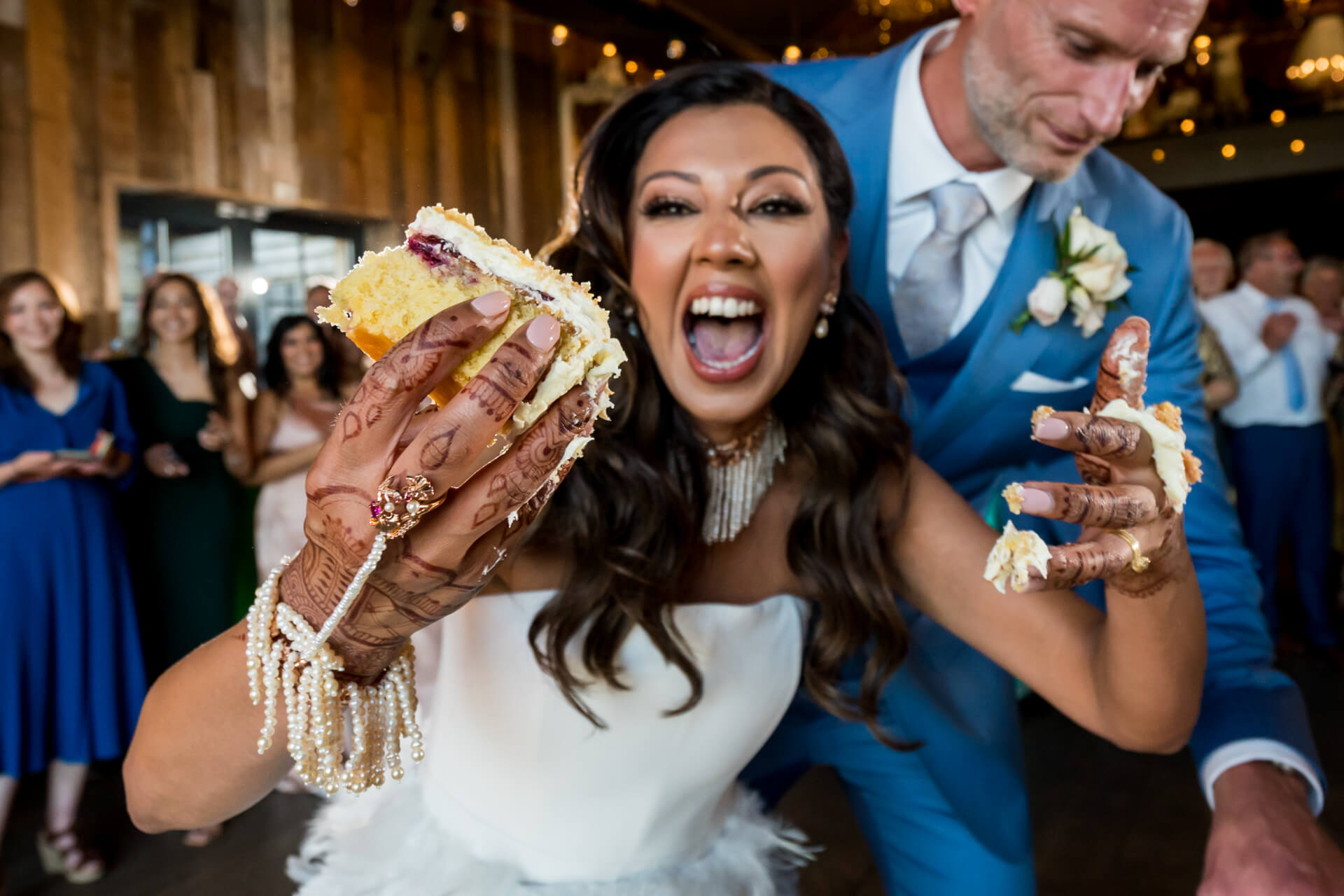 Bride joyfully smashing wedding cake with groom, guests smiling.
