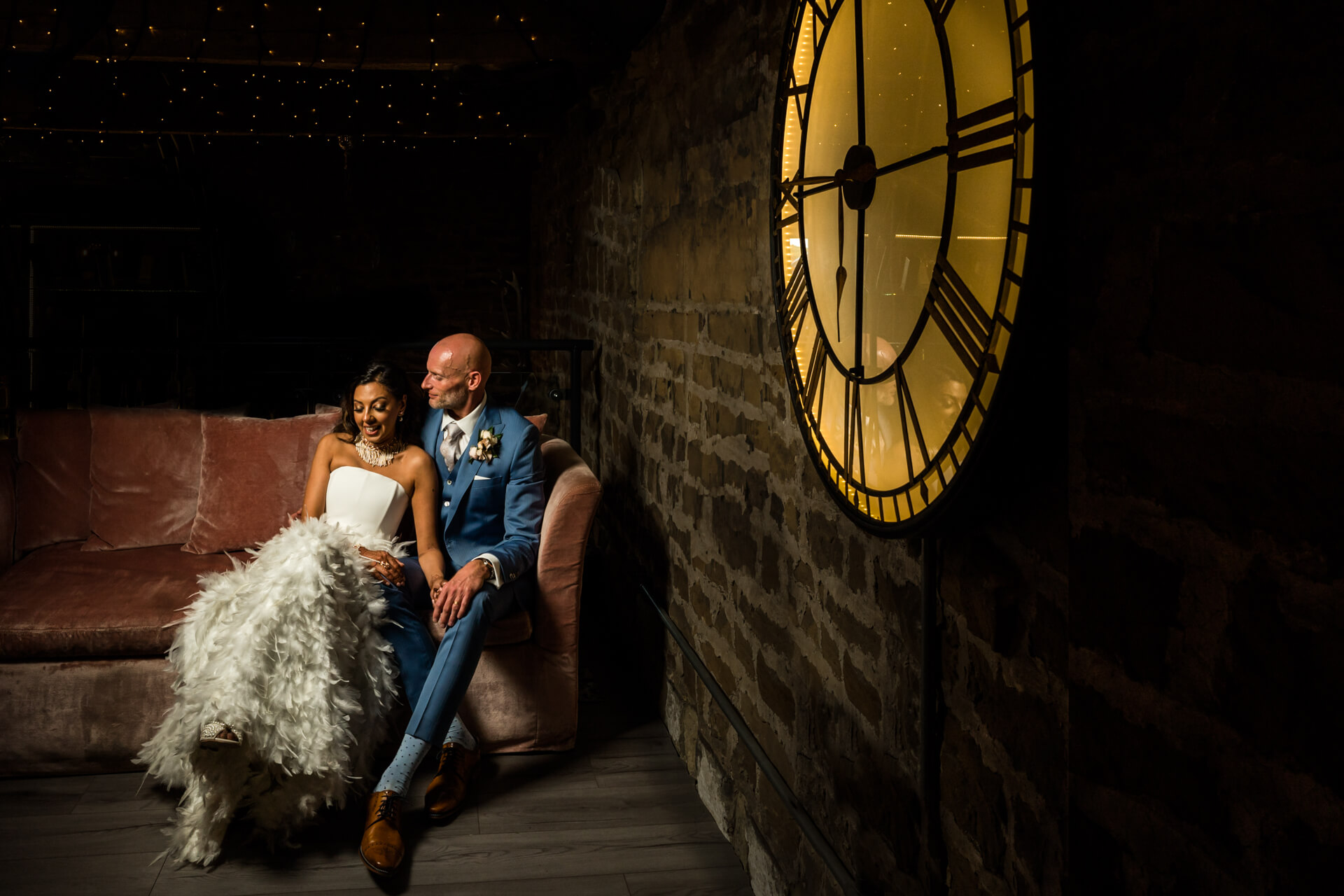 Wedding couple posing by large clock in ambient lighting.