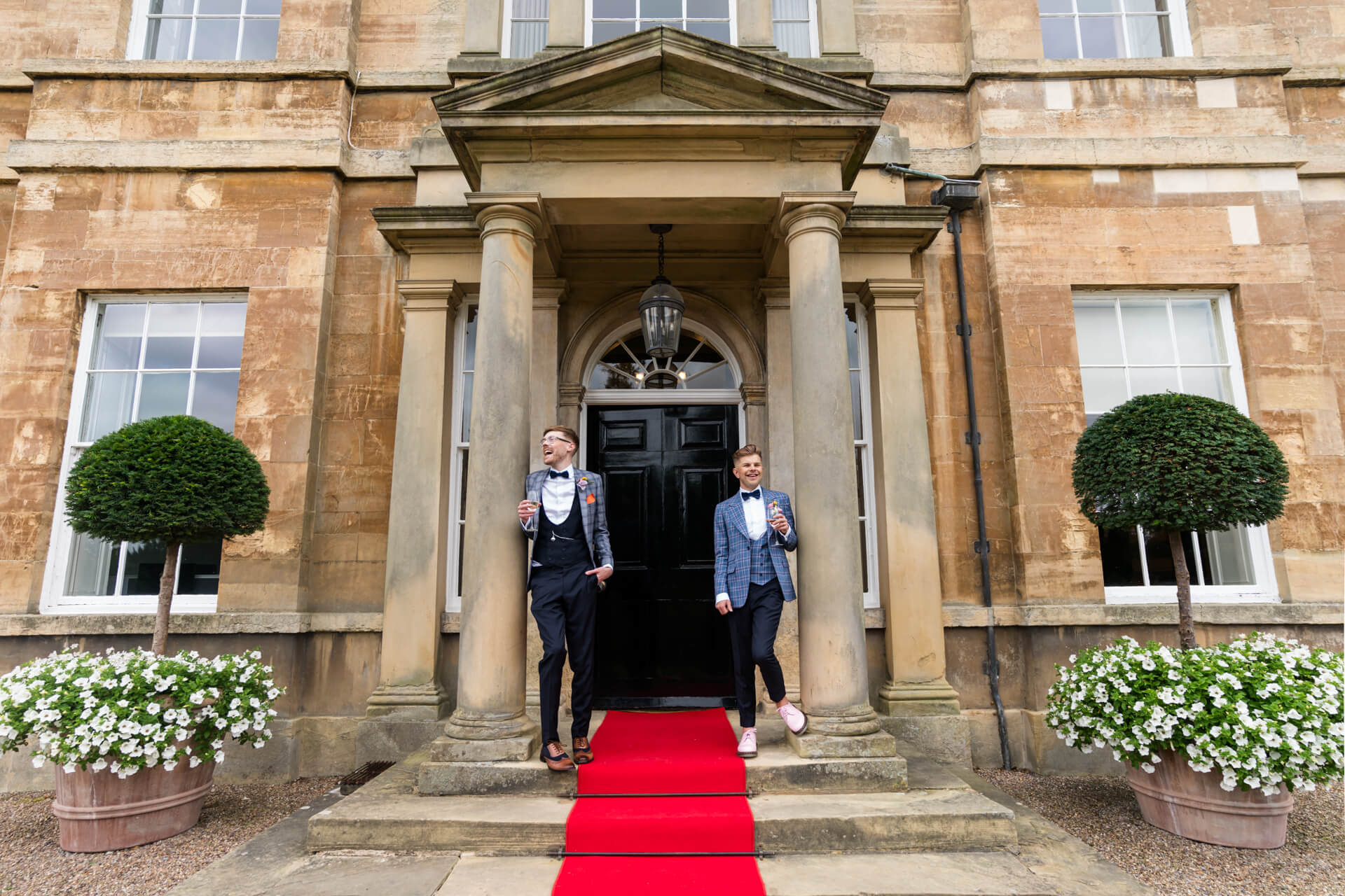 Groom couple standing on the front porch of Bowcliffe Hall holding champagne glasses
