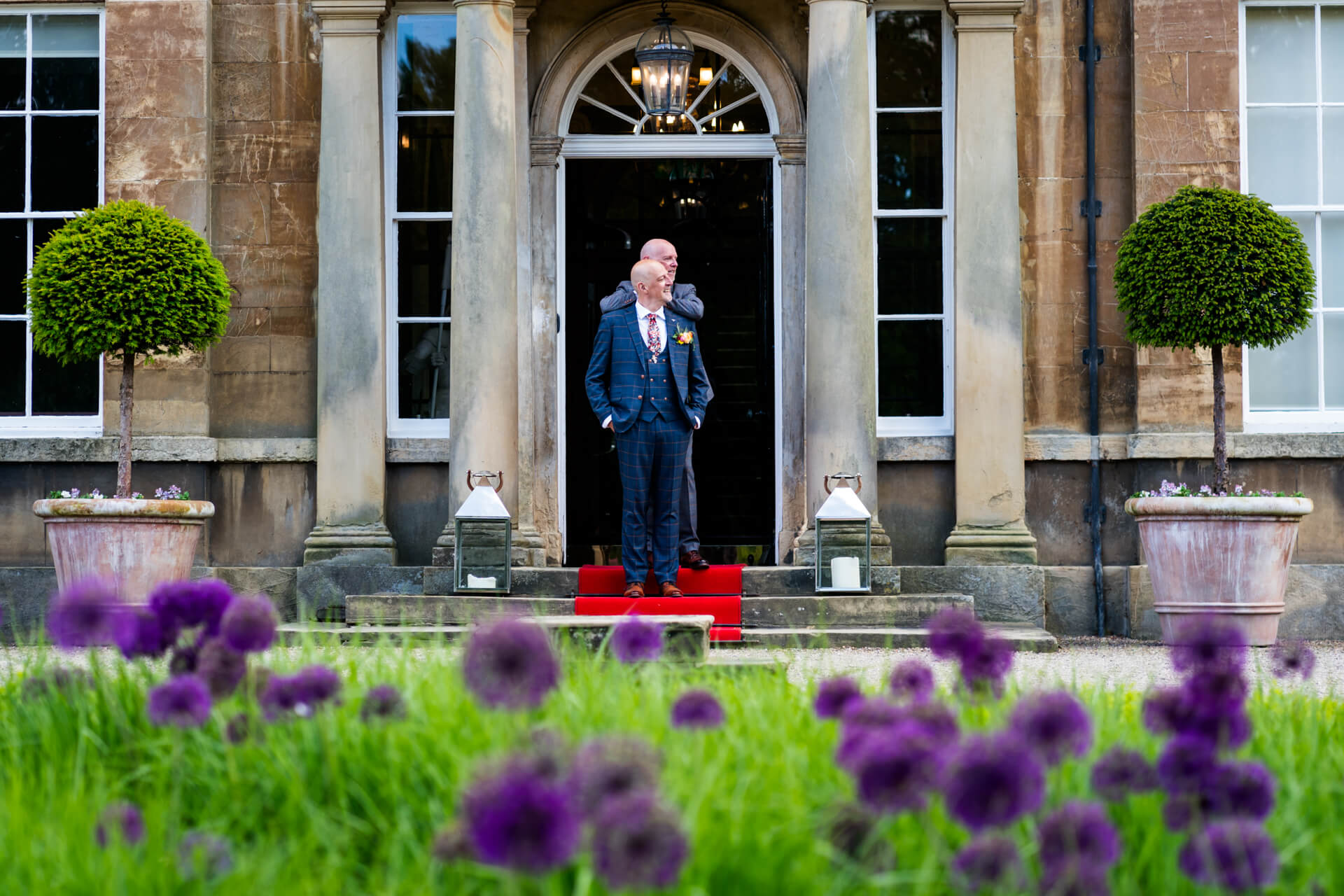 Two grooms in suits standing at Bowcliffe Hall entrance with flowers.