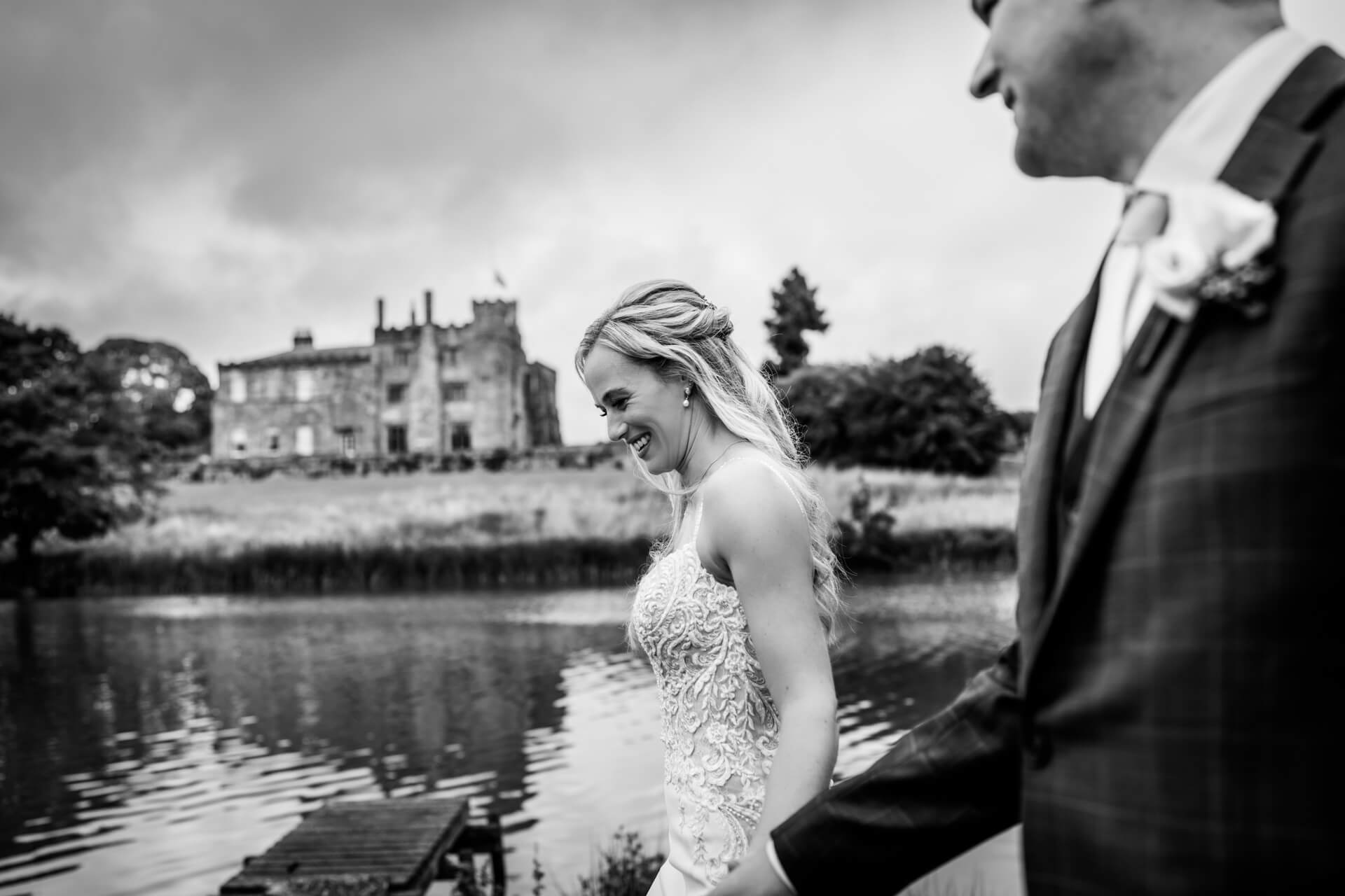 Bride smiling by lake, groom in foreground, Ripley castle background