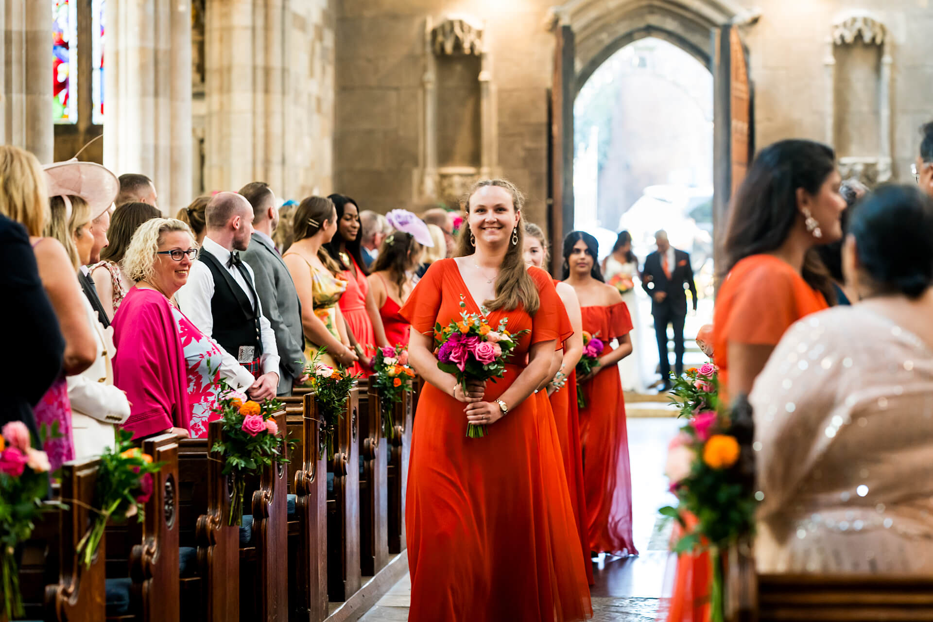 Bridesmaids walking down the aisle in orange dresses at church wedding ceremony.