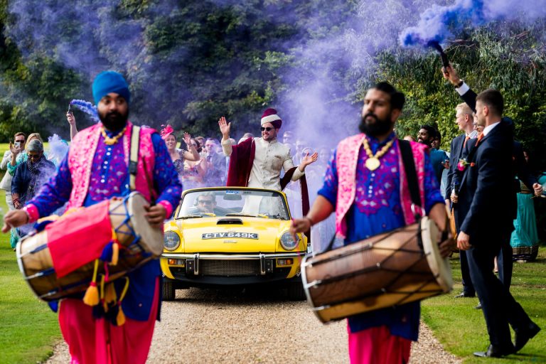 Colourful Indian wedding baraat at Rise Hall. Groom riding in a vintage car.