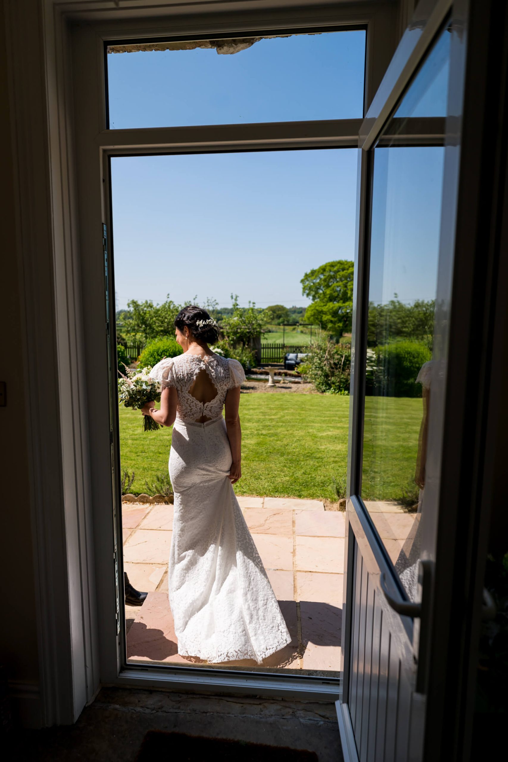 Bride in dress looking out at sunny garden.