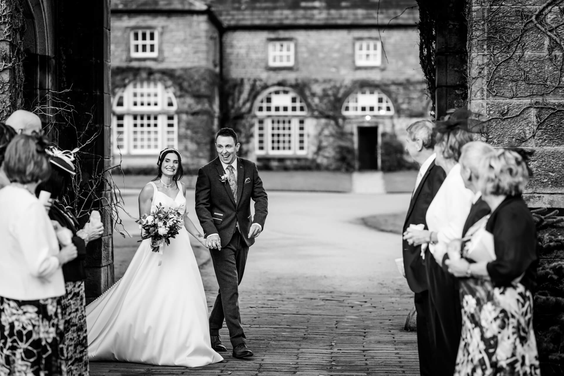 Bride and groom walking, guests clapping, historic building.