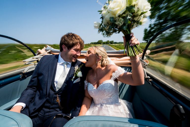 Bride and groom laughing in convertible car whilst its driving down the road outside the Whitehall Estate