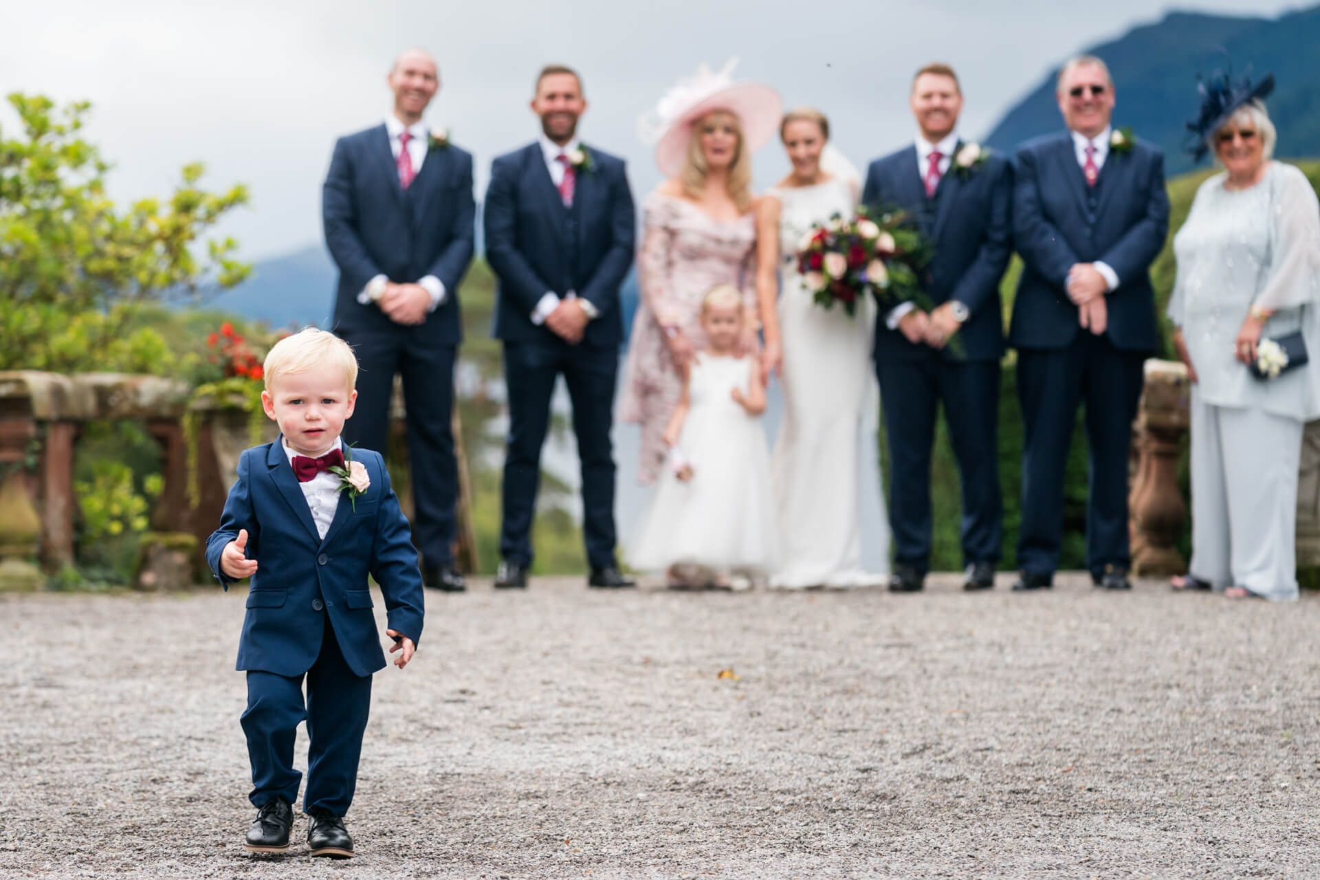 A young boy in a suit walking towards the camera with a wedding party blurred in the background, standing outdoors with mountains in the distance.