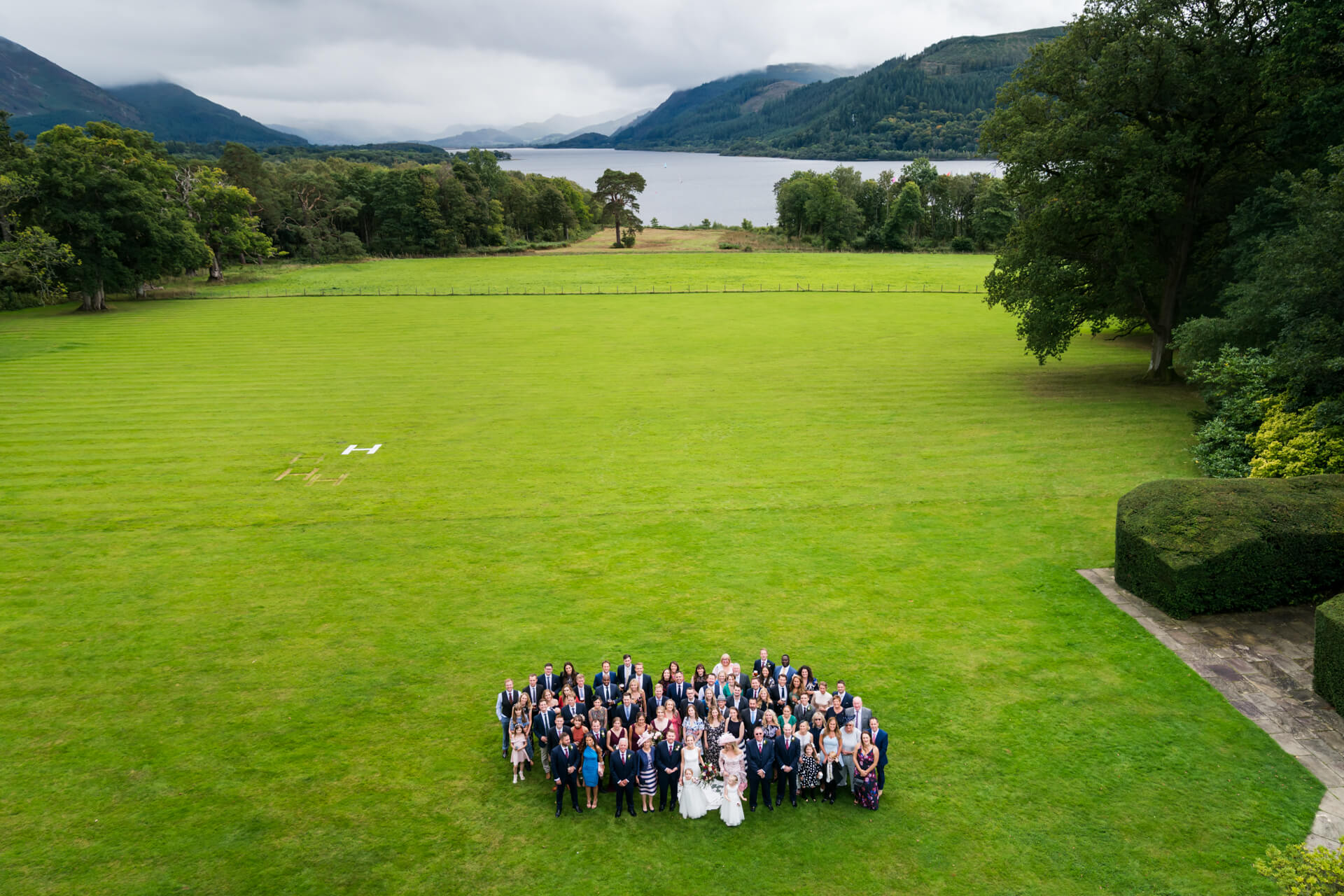 Aerial view of a large wedding group posing in a heart shape on a lush green lawn with a scenic lake and hills in the background.