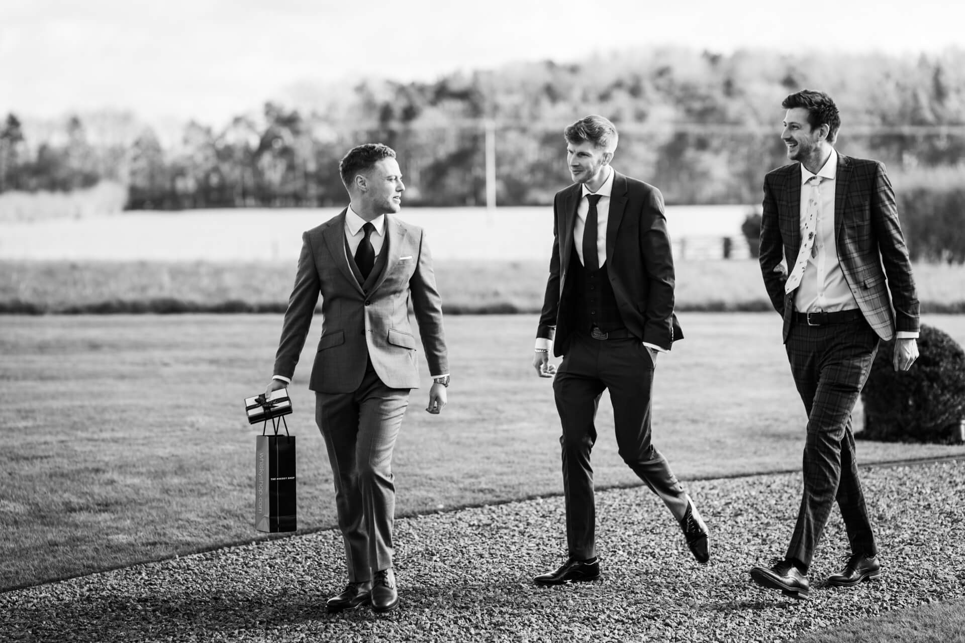 Groom and groomsmen in suits walking outdoors, smiling.