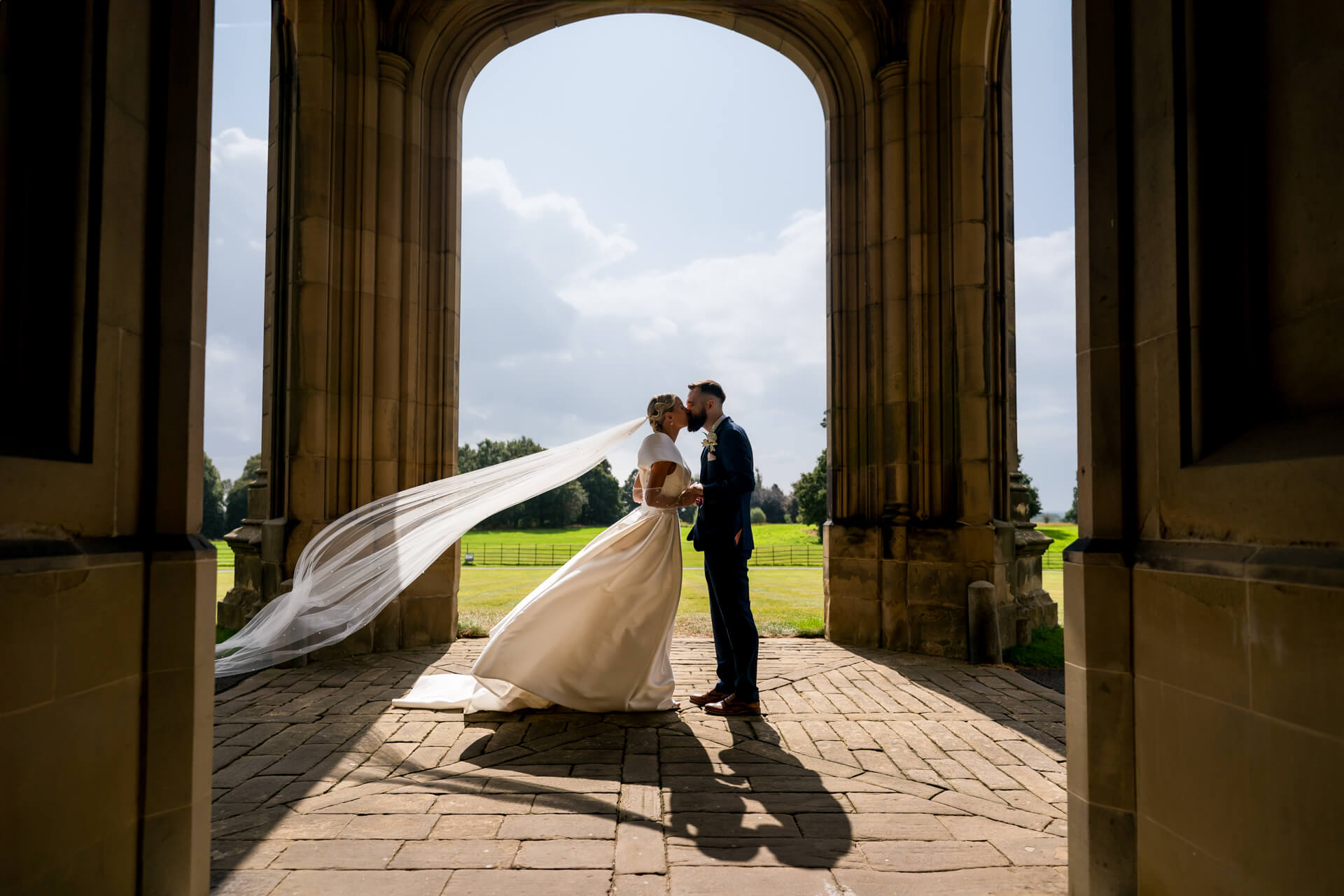 Bride and groom kissing under archway at Allerton Castle