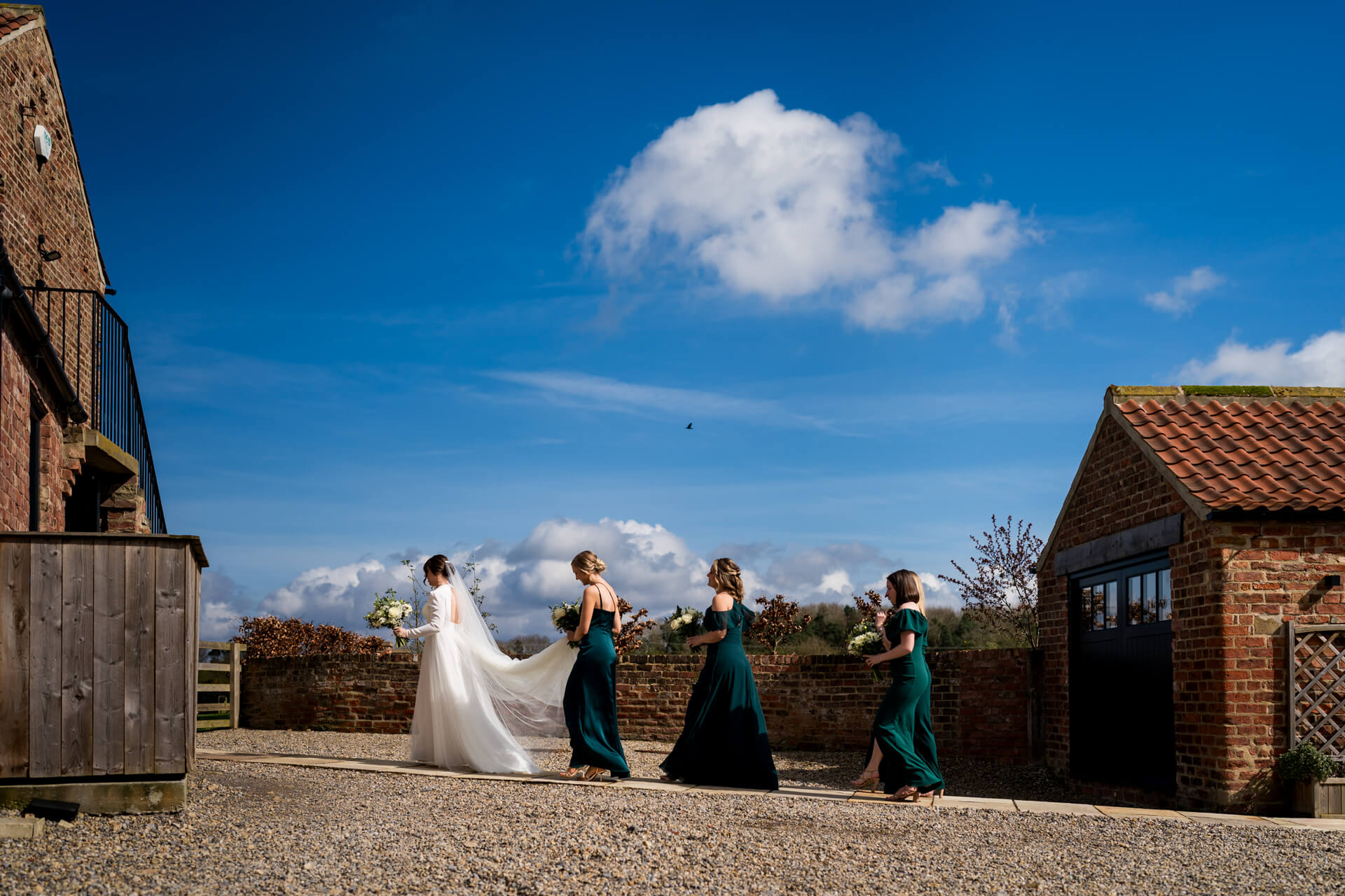 Bride and bridesmaids walking near brick buildings under blue sky at Thirsk Lodge barns