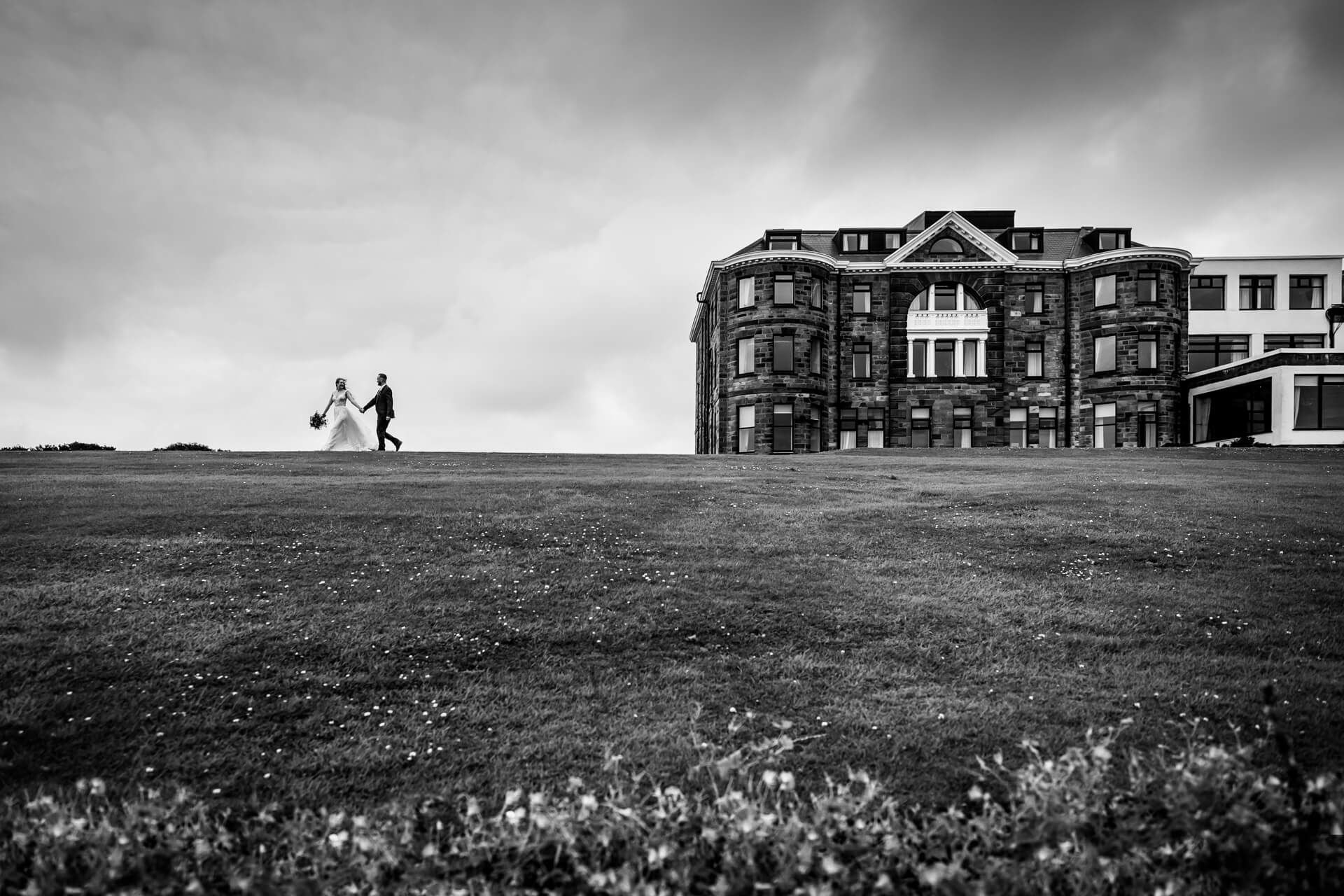 Bride and groom walking by historic Raven Hall hotel on grassy field