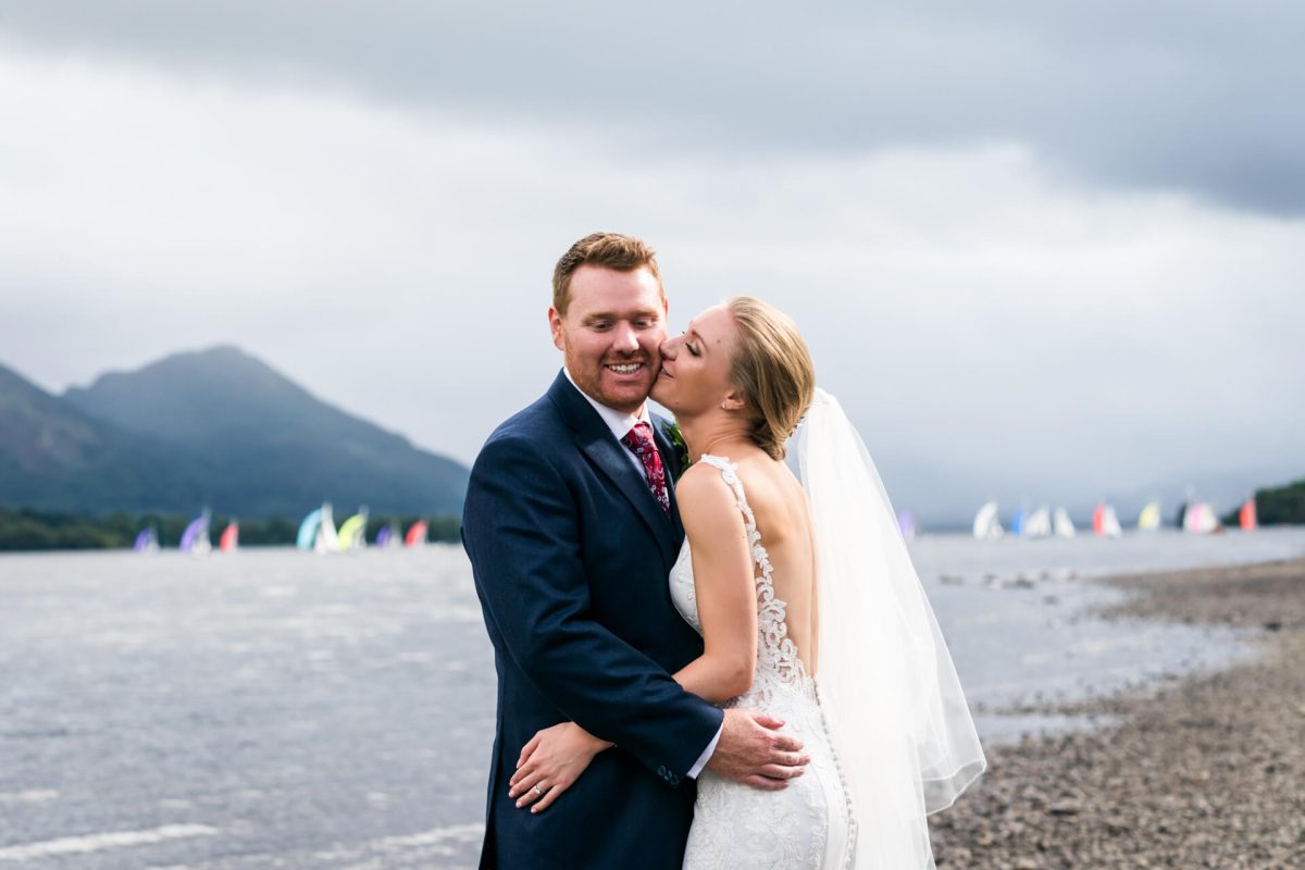 Bride kissing groom by lakeside with colorful sailboats