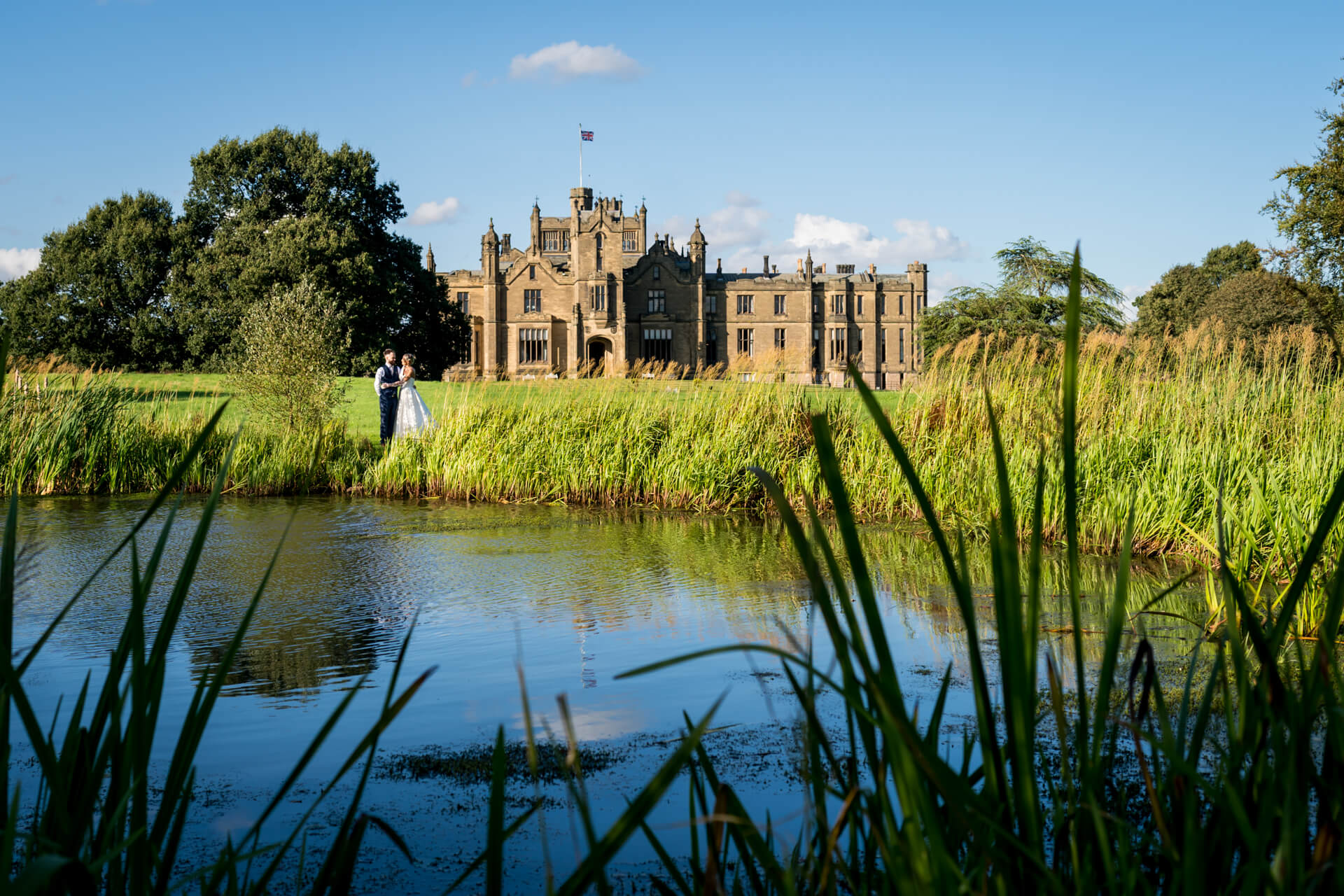 Wedding couple by lake in front of historic Allerton Castle on sunny day