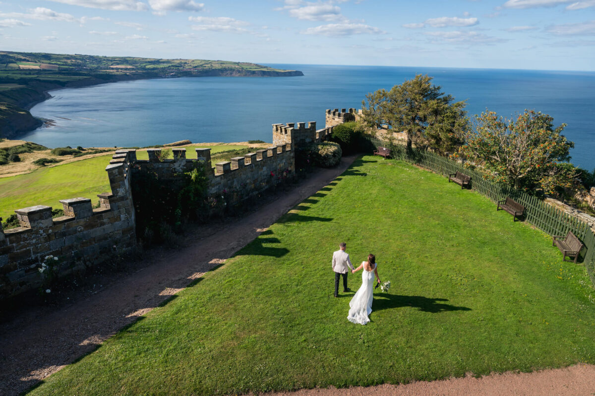 Couple walks on grass by seaside castle walls.